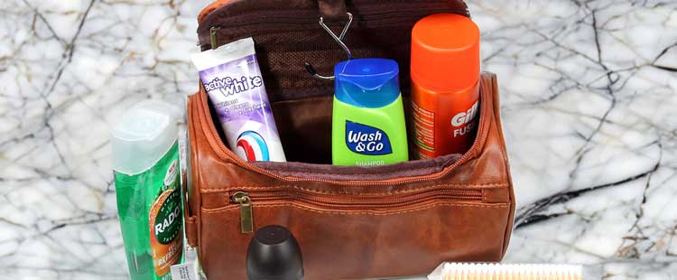 A brown toiletry bag open on a marble surface containing toothpaste, green shampoo, orange shaving cream, and a toothbrush. Additional items like a black deodorant and green Radia shampoo are nearby.