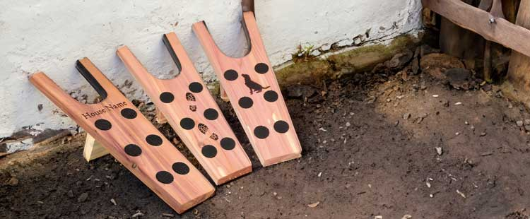 Three wooden wine holders with black dots are leaning against an outdoor wall. Each has unique engravings: one with House Nane, another with animals, and the third with a silhouette of a dog. Soil and a wooden fence are nearby.