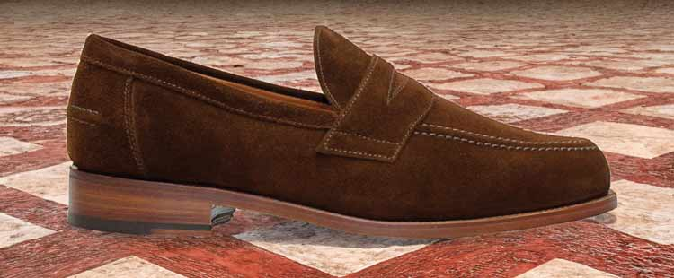 Brown suede loafer with a strap detail is displayed against a patterned tiled floor. The shoe has a wooden sole and minimal stitching.