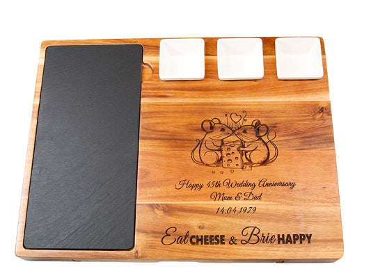 The Cathcart Elliot Deluxe Acacia Wood Cheeseboard Set (44x33cm) features an engraved message, three white dishes, a black slate meat platter, wire cutter, and two mice with cheese. Text: Eat cheese & brie happy plus your personalized anniversary message.
