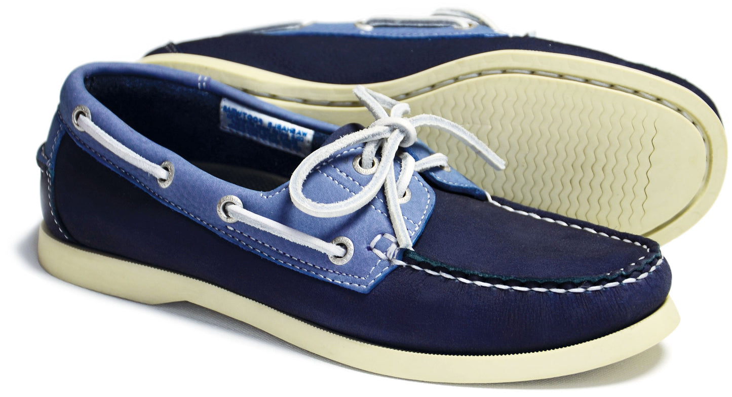 The SANDUSKY Ladies Indigo and Blue Deck Shoe by Orca Bay features white stitching, white laces, and cream non-slip soles. One shoe stands upright while the other is on its side showing the sole.