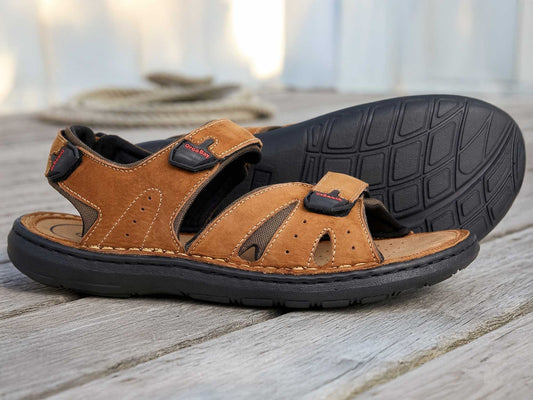The Orca Bay Tresco Brown Leather 3-Strap Sandal with a PU sole is displayed on a wooden surface, one men's sandal lying flat and the other propped to show its textured sole.