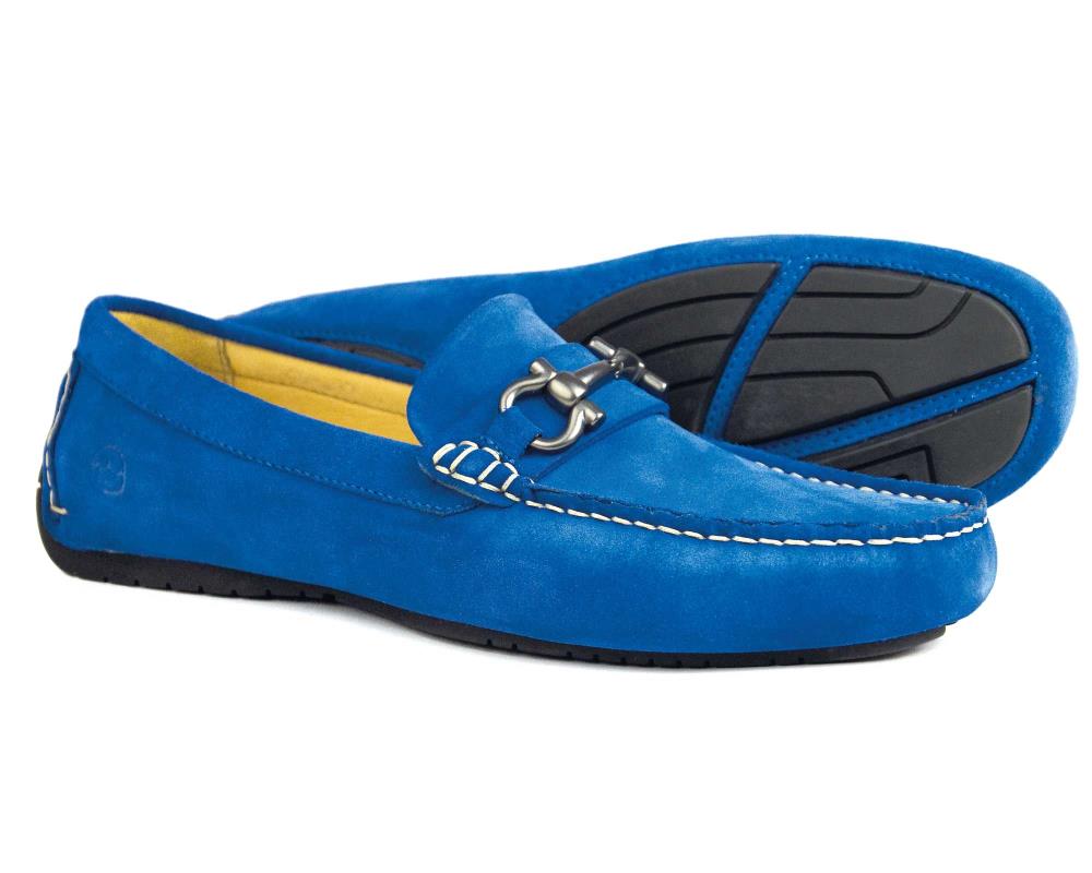 The ROMA 2 Men’s Royal Blue Suede Driving Loafer by Orca Bay features white stitching, black rubber soles, and a metal buckle on the front. One shoe lies flat; the other is positioned to display its sole.