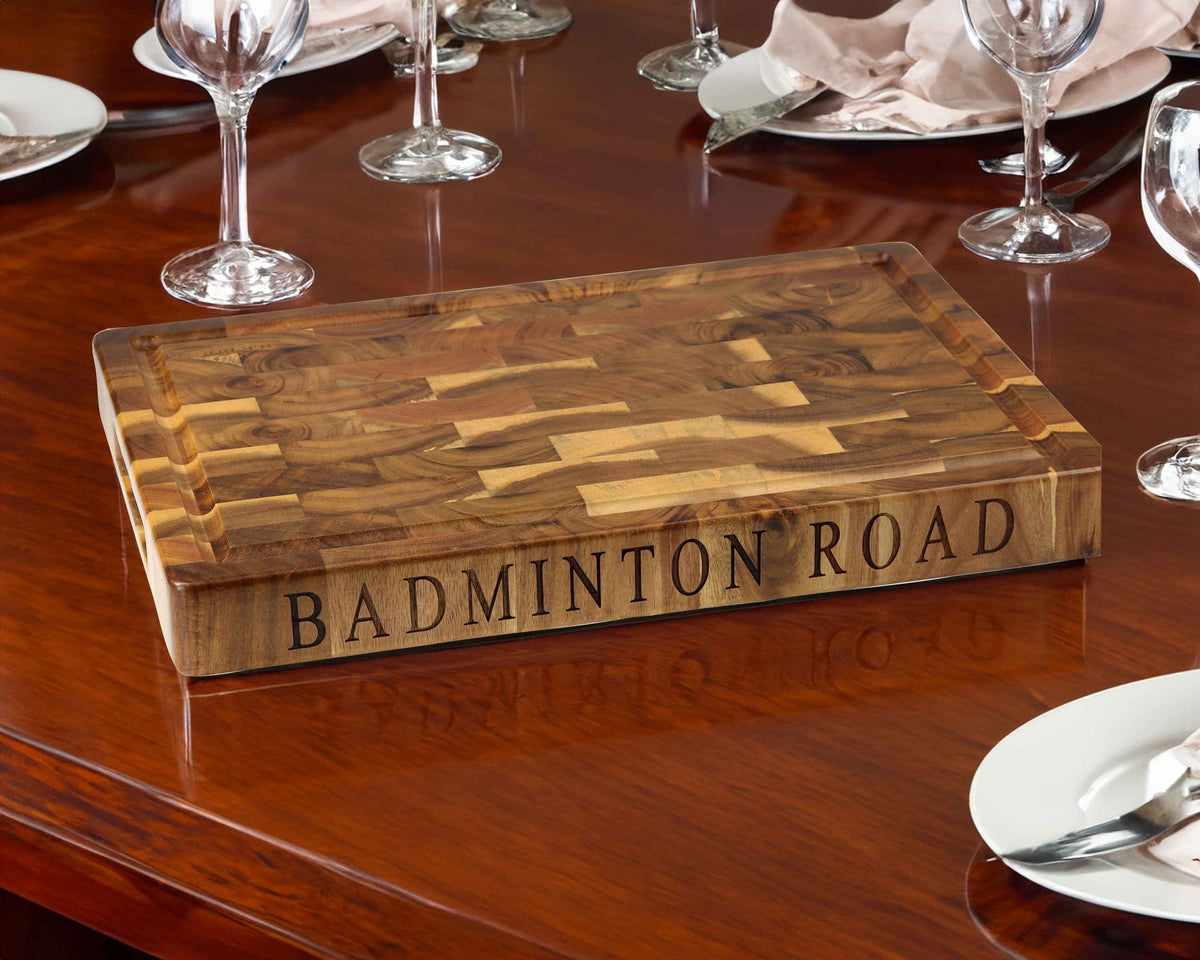 A Cathcart Elliot Large Acacia Wood Carving Chopping Cutting Board 40x30x4cm engraved with BADMINTON ROAD rests on a polished dining table, surrounded by plates, utensils, and glasses set for a meal.