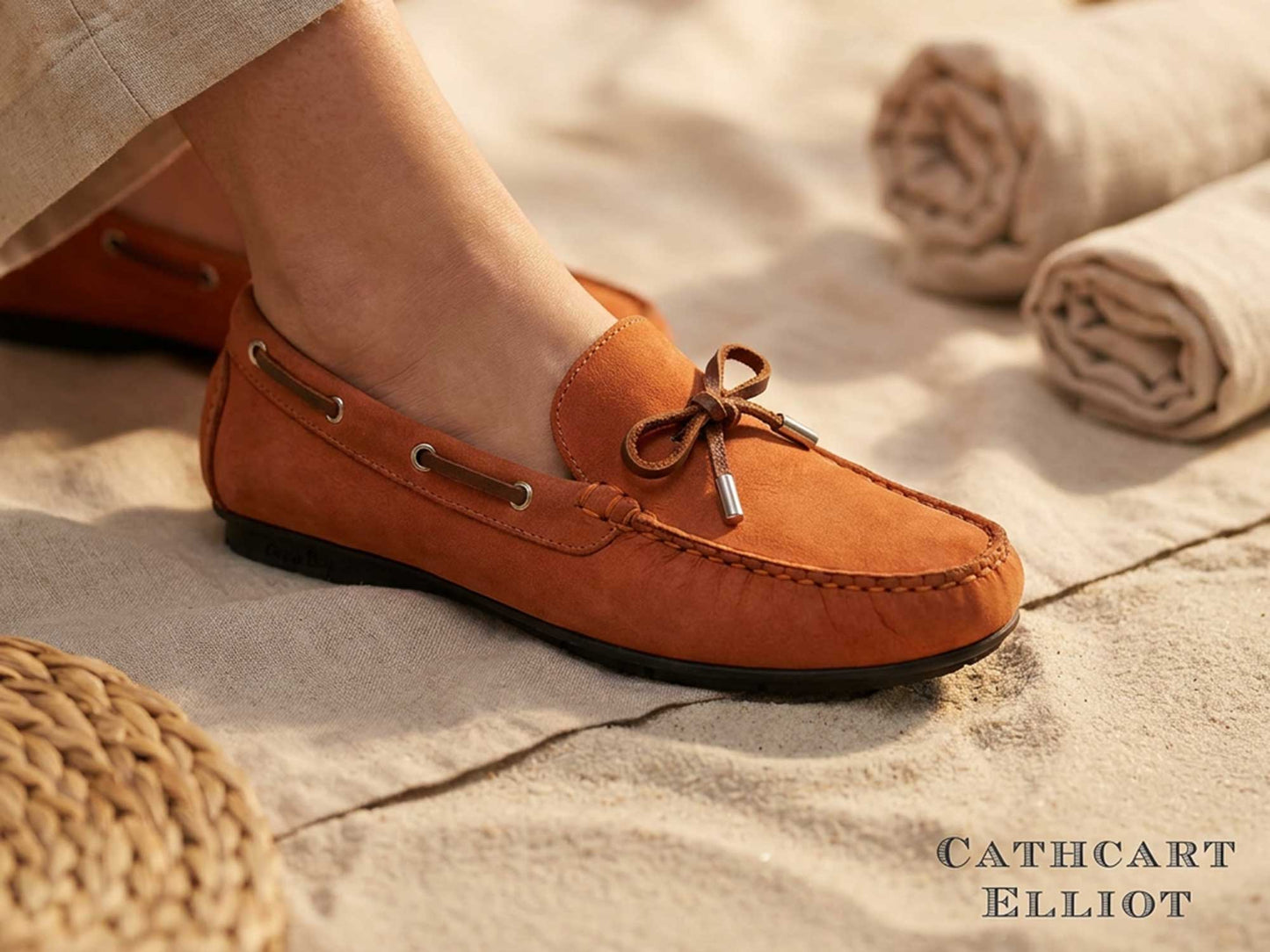 A close-up of a person wearing Orca Bay Ballena Ladies Burnt Orange Loafers with bow detail and moccasin construction, standing on sand near rolled towels and a woven mat. The text Orca Bay appears in the bottom right corner.