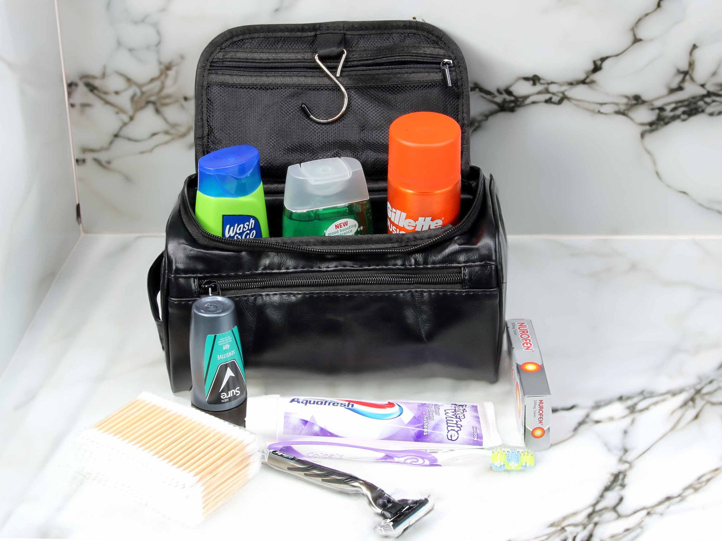 A Cathcart Elliot Black PU leather Washbag rests on a marble counter, containing deodorant, shaving gel, and shampoo. In front are a toothbrush, toothpaste, razor, comb, cotton swabs, and dental floss.