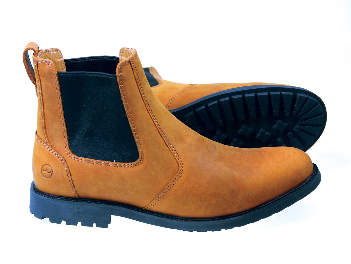 The Orca Bay BRECON Men’s Chelsea Boots in Sand Nubuck feature black elastic side panels, thick black rubber soles, and a water-resistant finish for durability—one boot upright, the other on its side displaying the tread.