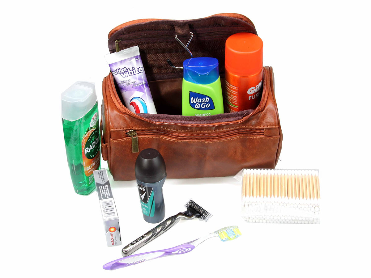 The Cathcart Elliot Brown PU leather Water Resistant Washbag with Hanger Hook is shown open with shampoo, toothpaste, and deodorant inside. In front of the bag are a toothbrush, razor, mouthwash, shoe polish, cotton swabs, and a deodorant stick.