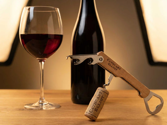 A glass of red wine, a wine bottle, and the Cathcart Elliot Personalised Wine Corkscrew and Bottle Opener with a beech wood handle holding a cork, all placed on a wooden surface with gentle lighting in the background.
