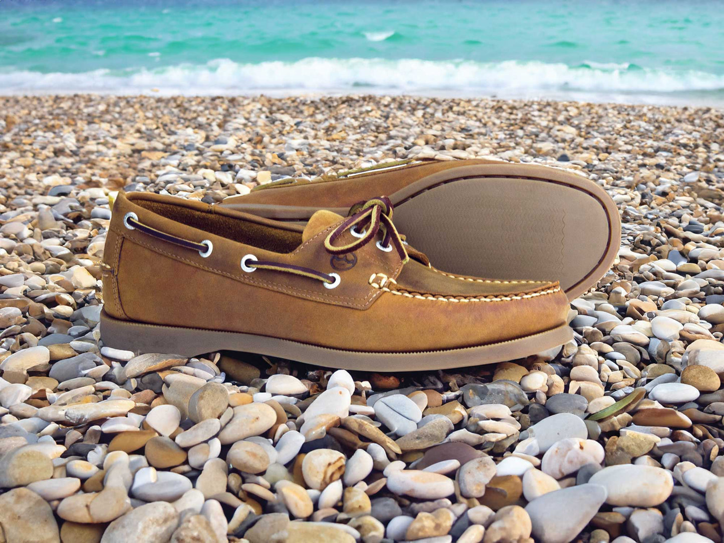 The Women’s Boat Shoes | Orca Bay Creek Sand Nubuck by Orca Bay sit on a pebbly beach beside turquoise waves under a clear sky.