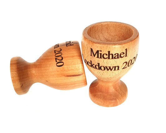 The Cathcart Elliot Personalised wooden egg cup set of 2 features beech wood cups, one upright engraved with Michael Lockdown 2020 and another on its side, both on a plain white background.