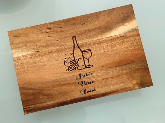 The Cathcart Elliot Personalisable Rustic Large Acacia Wood Cheeseboard Set showcases an engraved wine bottle, glass, cheese, grapes, and Josies Cheese Board in a stylish font. Its rich natural grain makes it a perfect gift for cheese lovers. Size: 36cm x 24cm.