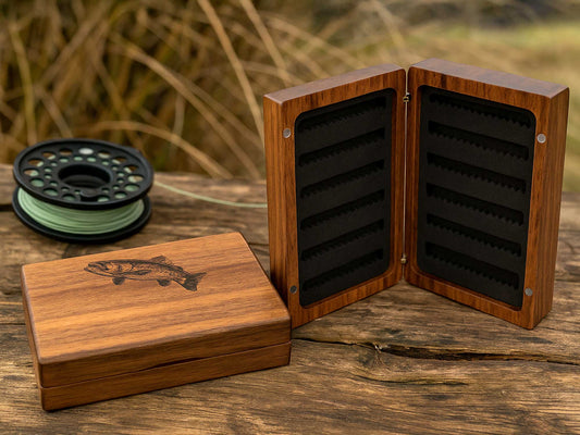 The Personalised Pocket Sized Walnut Fishing Fly Box by Cathcart Elliot, featuring a fish engraving, sits open on rustic wood—its lid beside it and a green-line fishing reel in the background—making it an ideal gift for anglers.