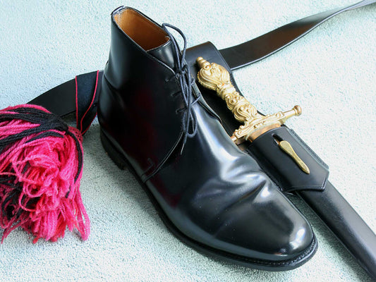 A shiny black dress shoe, a black leather belt with a gold-handled ceremonial dagger in its sheath, and a red and black tassel lay on a light-colored surface. Beside them rests the Sanders George 5634B Black Military Dress Boot by Sanders—crafted with Goodyear welted construction—completing the distinguished arrangement.