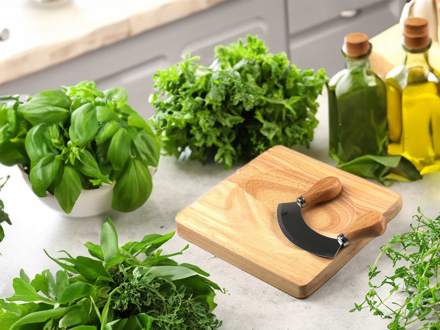 A Cathcart Elliot mezzaluna knife sits on a Personalised mezzaluna wooden herb chopper board, surrounded by basil and parsley. In the background, two olive oil bottles elegantly adorn the kitchen counter.
