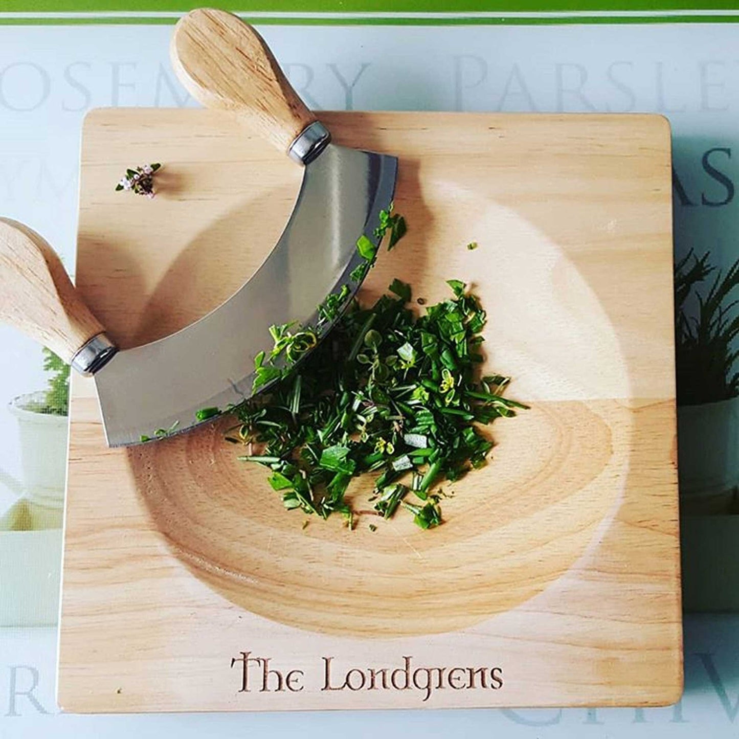 A mezzaluna knife with wooden handles sits on a Cathcart Elliot herb chopping board. Chopped green herbs are in the center. The personalized rubber wood board, ideal for any stainless steel chopper, is engraved with The Londgrens.