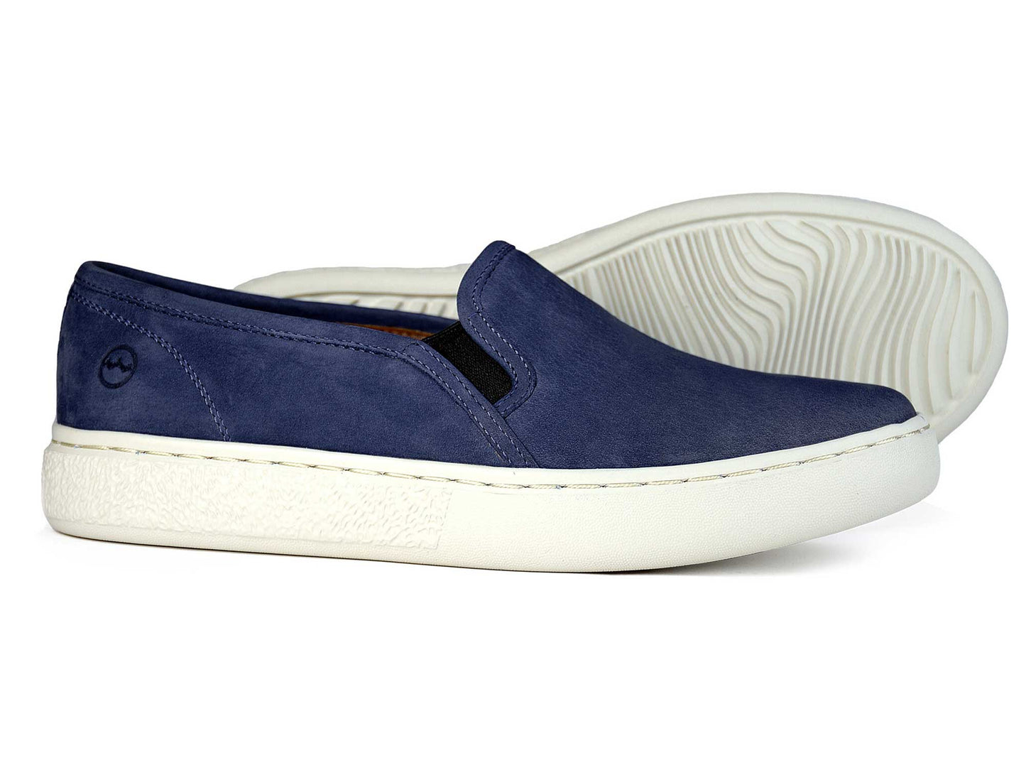 A pair of Orca Bay MARYLEBONE Ladies Indigo Blue Summer Loafers feature soft nubuck leather and white textured rubber soles. One slip-on shoe stands upright; the other lies on its side, revealing the sole’s pattern.