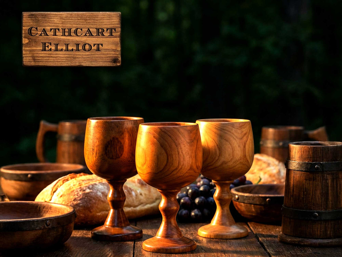 Three Cathcart Elliot Engraved Jujube Wood Wine Goblets rest on a rustic table with bread, grapes, bowls, and tankards beneath a wooden CATHCART ELLIOT sign—set against a dark forest, the ideal unique wine gift.
