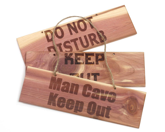 Three Cathcart Elliot cedar wood door plaques (20x6 cm) overlap: a custom-engraved DO NOT DISTURB, a partially visible KEEP OUT, and a rope-hung Man Cave Keep Out—all in bold, dark lettering.
