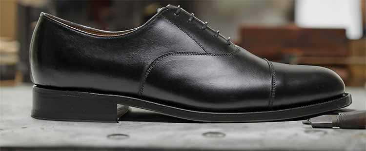 A polished black leather oxford shoe with a sleek design is displayed on a smooth surface. The shoe features a cap toe and appears elegant, suitable for formal occasions.