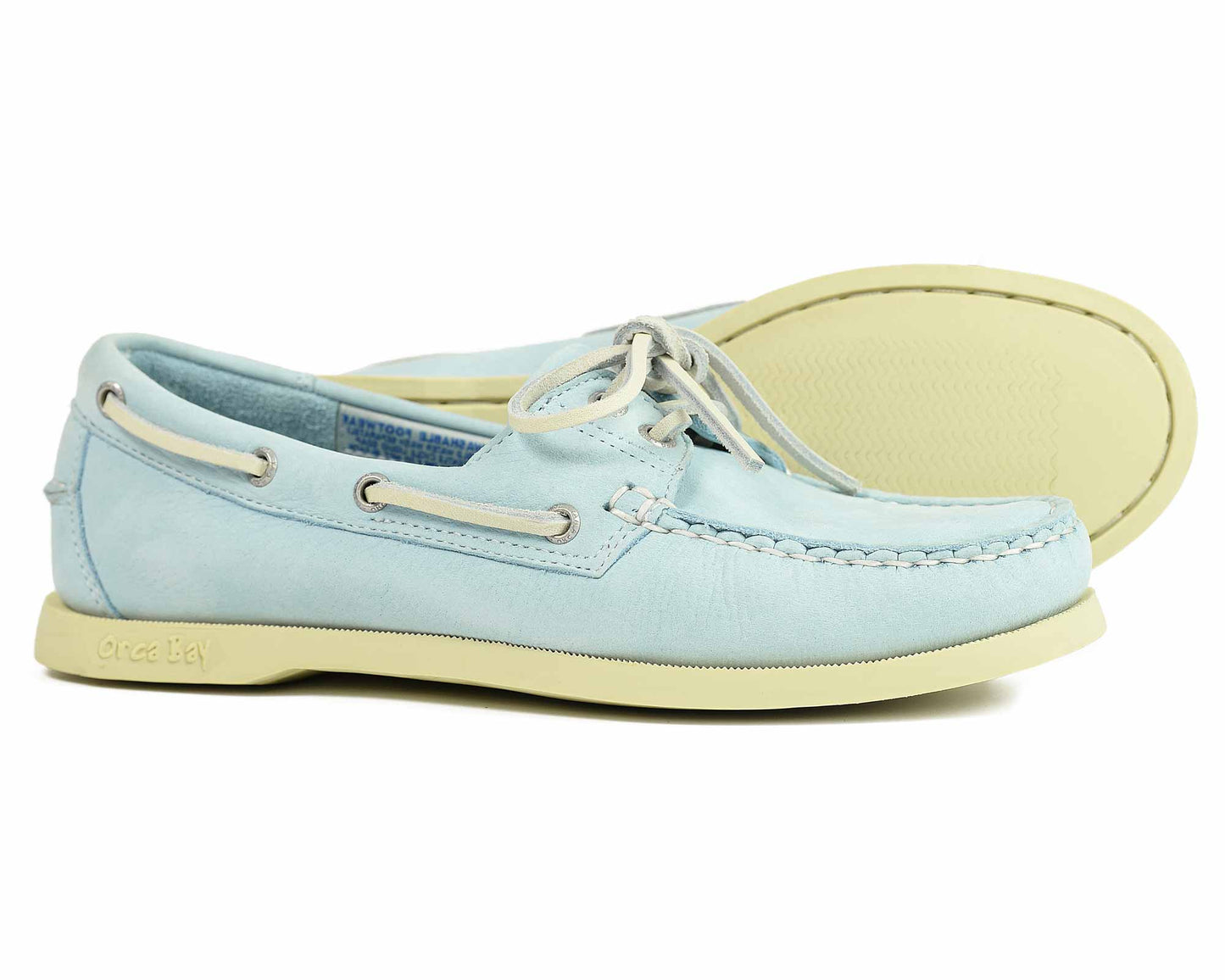 The Orca Bay SANDUSKY Ladies Boat Shoes in Ice Blue feature cream-colored non-slip soles, white laces, metal eyelets, visible stitching, and an embossed logo on the outer heel. One shoe is upright while the other tilts on its side.