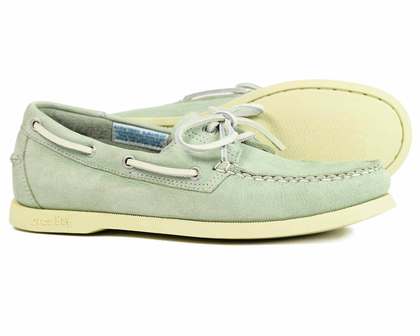The SANDUSKY Ladies Boat Shoes by Orca Bay are sage green suede with white laces and cream-colored soles. They feature decorative stitching and metal eyelets. One shoe stands upright, while the other displays its non-slip sole with an intricate tread pattern.