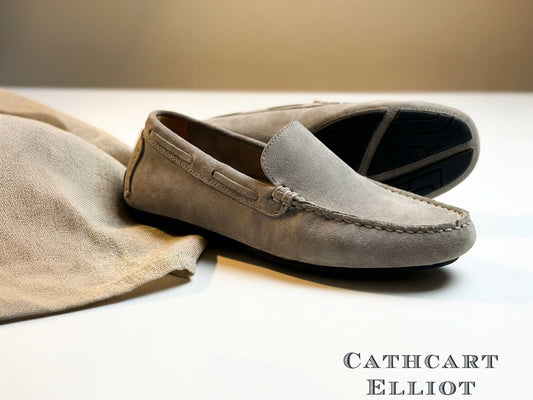 The Orca Bay Silverstone Men's Grey Suede Driving Loafers are displayed on a white surface next to a matching suede cloth, with the Orca Bay brand name shown in the bottom right corner.