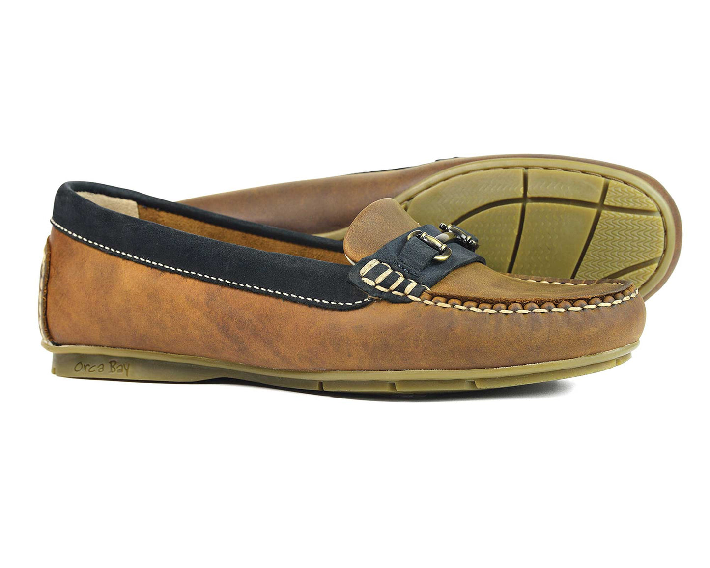 The VERONA Ladies Sand & Navy Buckled Loafer by Orca Bay features elegant sand and navy leather with a buckle detail. One loafer stands upright, while the other rests on its side, showcasing their design and unique sole accents.