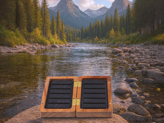 The Cathcart Elliot Personalised Pocket Sized Walnut Fishing Fly Box (14x9cm) sits open on river rocks amid pines and mountains—a thoughtful gift for anglers.