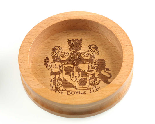 The Cathcart Elliot Personalised Wine Bottle Coaster with Crest features a round wooden design engraved with your family crest—shield, lion, figure with branch—and the name “BOYLE” at the bottom.