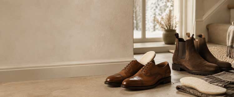 A pair of brown dress shoes and brown boots are placed neatly on a beige floor near a window and stairs, with shoe inserts resting beside them. A potted plant sits on the windowsill.