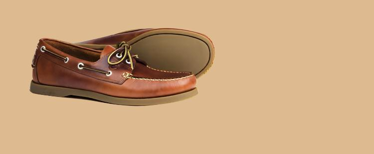 Go to the A pair of brown leather boat shoes with laces and white stitching, placed against a solid beige background. The shoes have a non-slip sole and metal eyelets. One shoe is upright while the other is on its side. collection