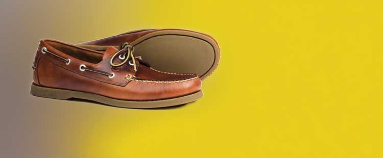 A pair of brown leather boat shoes with white stitching and lace details is displayed on a gradient yellow to brown background. One shoe is upright, while the other leans on its side, showcasing the sole.