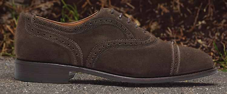Brown suede brogue shoe with detailed perforations, showcased on a gray surface with a blurred dark background featuring some green grass.