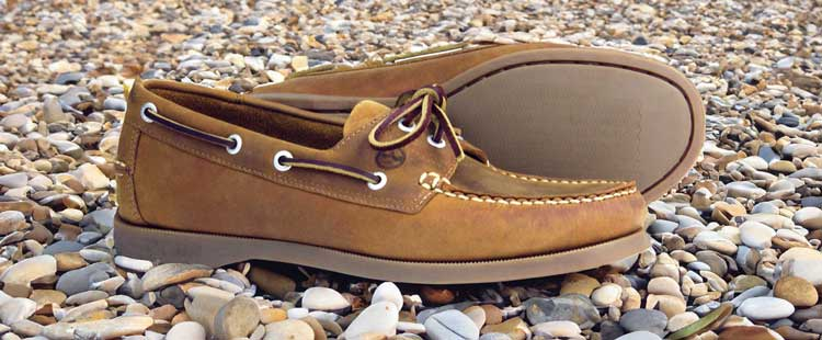 Brown boat shoes with white laces and stitching, placed on a pebble-covered ground. One shoe lies on its side, displaying the sole.