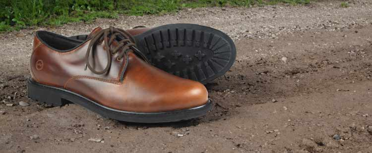 A pair of brown leather shoes with black soles placed on a muddy, gravel path. The shoes have laces and one shoe is on its side, showing the textured sole.