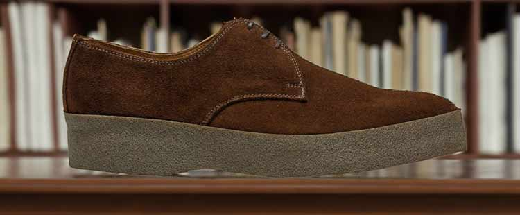 Brown suede shoe with thick gray sole on a wooden surface, background features blurred bookshelf.
