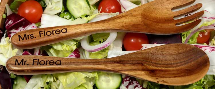Wooden salad tongs labeled Mr. Florea and Mrs. Florea rest on a bed of fresh salad ingredients, including lettuce, cherry tomatoes, cucumber slices, and red onion.