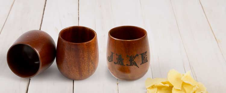 Three wooden cups are on a light wooden surface. Two cups are upright, one on its side. The upright cup on the right has the name JAKE engraved. A small pile of potato chips is nearby.