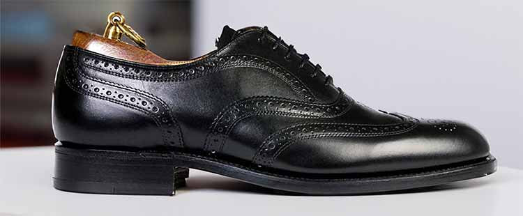 A polished black leather brogue shoe with detailed perforations and a wooden shoe stretcher inside, placed on a white surface.