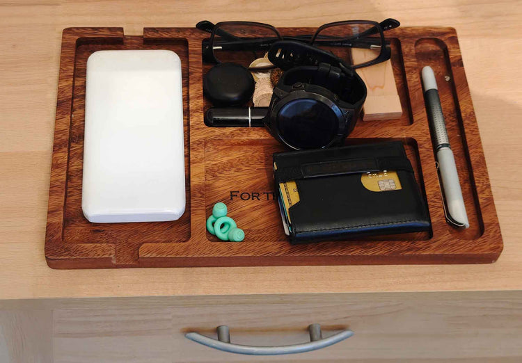 A wooden tray on a desk holds a white smartphone, glasses, a black watch, pen, wallet with a visible card, earplugs, lip balm, and a small black case.