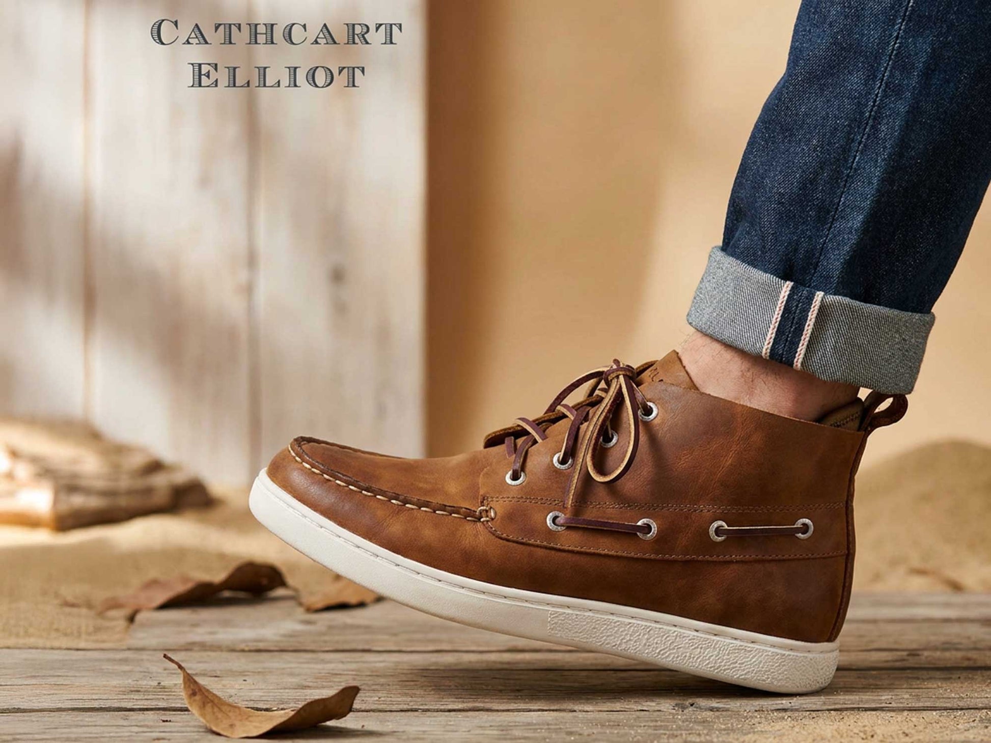 A person in rolled-up jeans and Orca Bay Annapolis Men’s Sand Oiled Nubuck Boots stands on a wooden surface strewn with dry leaves, with the Orca Bay logo visible in the background.