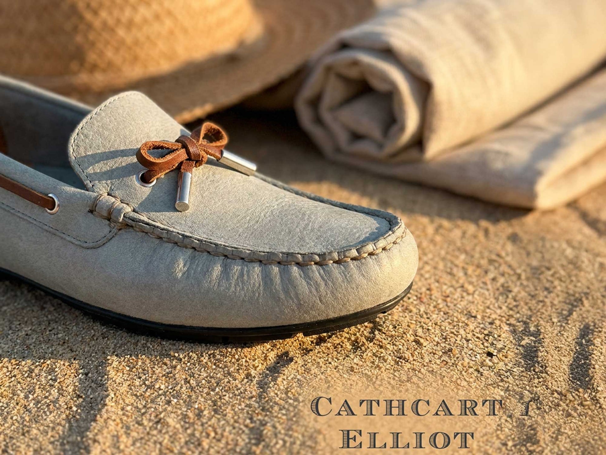 Close-up of the Orca Bay Ladies Ballena Pale Grey Washable Nubuck Loafer Shoe with brown lace bow on sand, next to a rolled-up beige cloth and straw hat. "CATHCART ELLIOT" text appears in the bottom right corner.