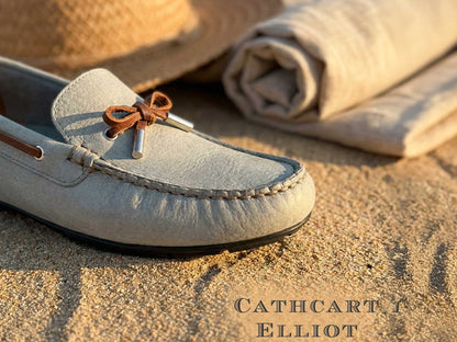 Close-up of the Orca Bay Ladies Ballena Pale Grey Washable Nubuck Loafer Shoe with brown lace bow on sand, next to a rolled-up beige cloth and straw hat. "CATHCART ELLIOT" text appears in the bottom right corner.