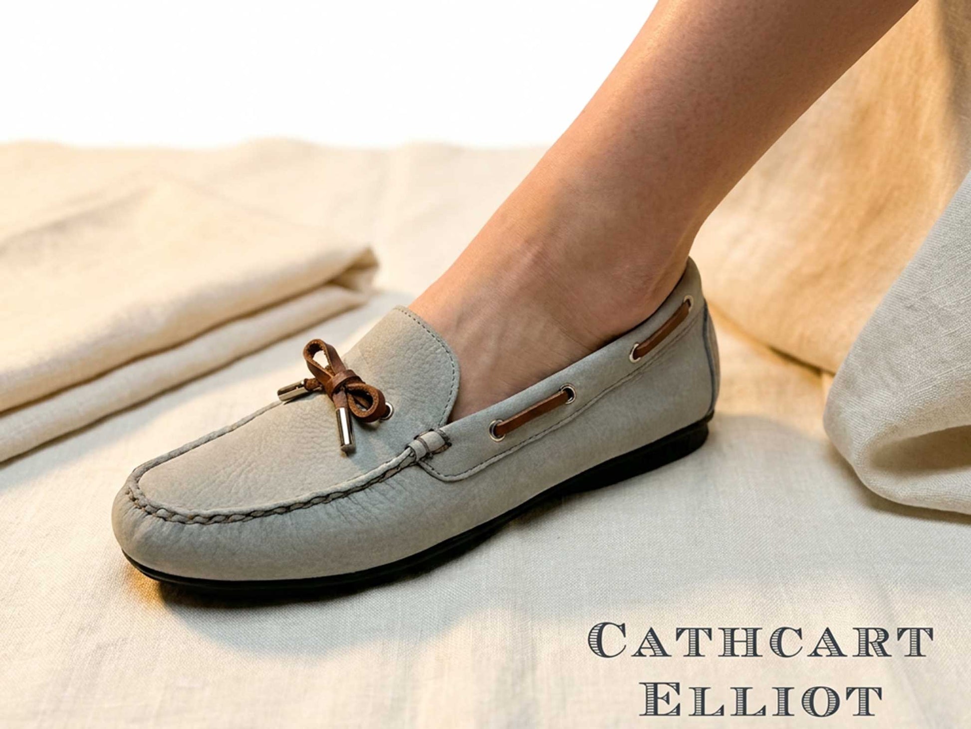 A close-up shows a person wearing the Ladies Ballena Pale Grey Washable Nubuck Loafer Shoe by Orca Bay, featuring brown leather laces. The shoe rests on light fabric, with "CATHCART ELLIOT" text in the corner—ideal for Orca Bay fans.