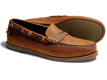 A pair of Orca Bay Men's Slip-On Deck Shoes in sand nubuck with visible stitching, moccasin construction, metal eyelets, and brown rubber soles. One shoe stands upright; the other lies on its side to show the sole.