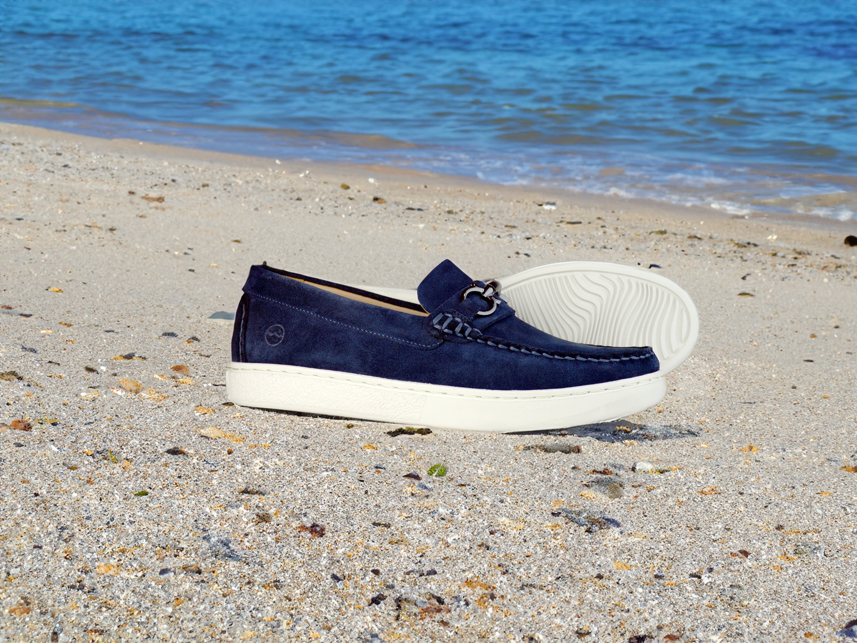 A pair of blue loafers with white soles is placed on a sandy beach, with the ocean and waves in the background.