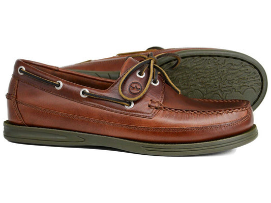 The Orca Bay FOWEY Men's Wide Fit Deck Shoe in elk brown leather features tan stitching, white eyelets, lace detail, and green slip-resistant soles. One shoe stands upright; the other lies on its side to display the textured sole.