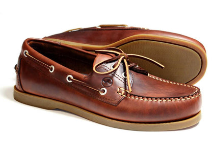 The Orca Bay Creek Men's Deck Shoe in saddle brown features salt-resistant leather, white stitching, tan rubber soles, and leather laces. One shoe stands upright while the other lies sideways, showing off the detailed tread.