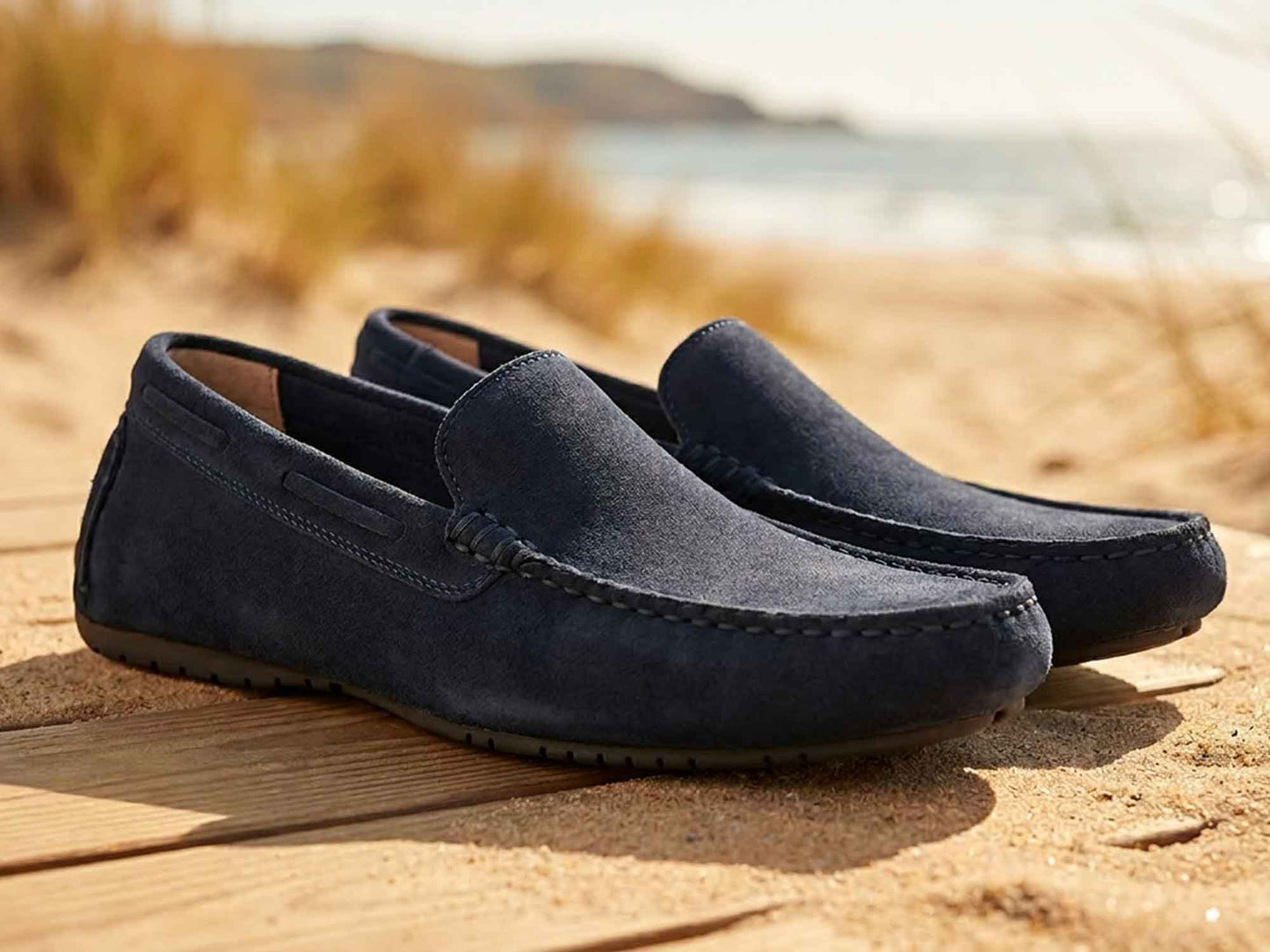 A pair of Orca Bay Silverstone Navy Suede Driving Shoes for men, featuring a non-slip rubber sole, rests on a wooden boardwalk with sand and a blurred beach backdrop.